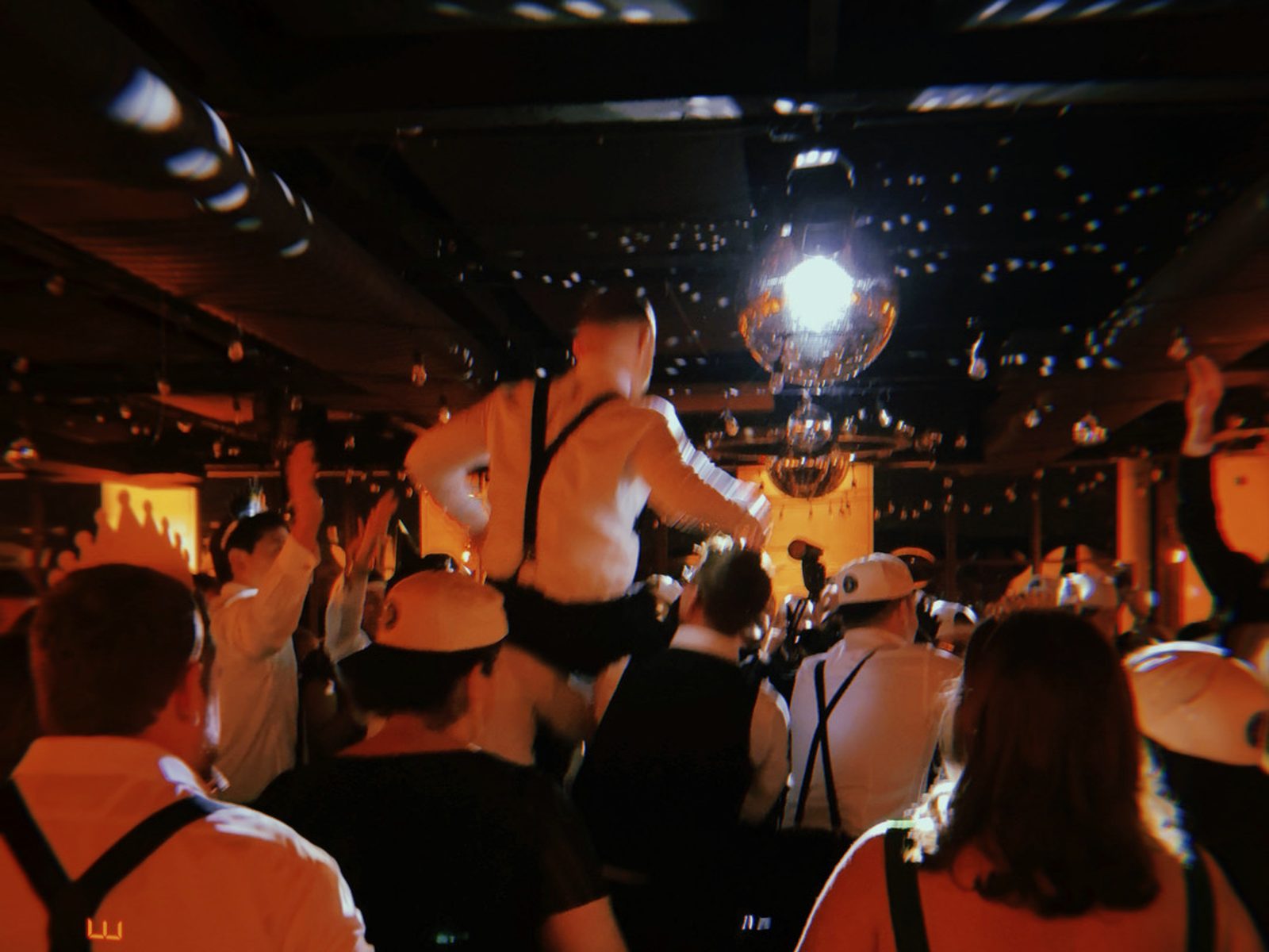 View from the DJ booth of a packed wedding dance floor under disco balls, with guests moving through a dark room.