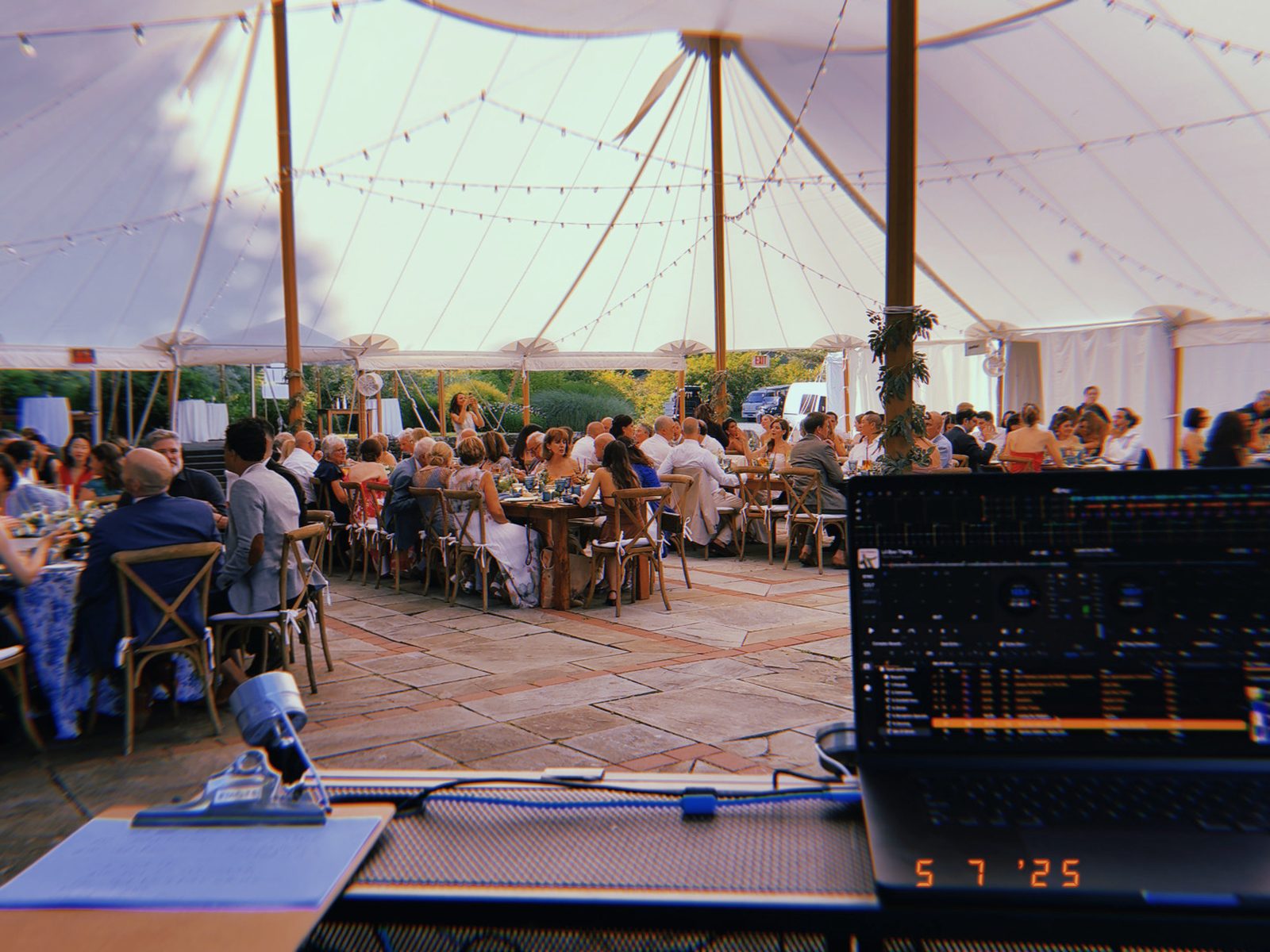 View from the DJ booth across a tented wedding dinner, with the controller, microphone, and room visible before dancing begins.